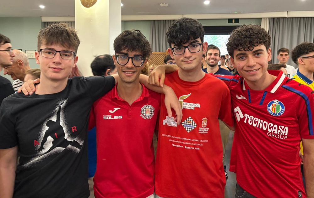 Cuatro jóvenes permanecen de pie, abrazados, con gafas y camisetas de temática deportiva, celebrando el Campeonato de España de ajedrez relámpago. Al fondo hay más gente, algunos sonriendo y hablando.