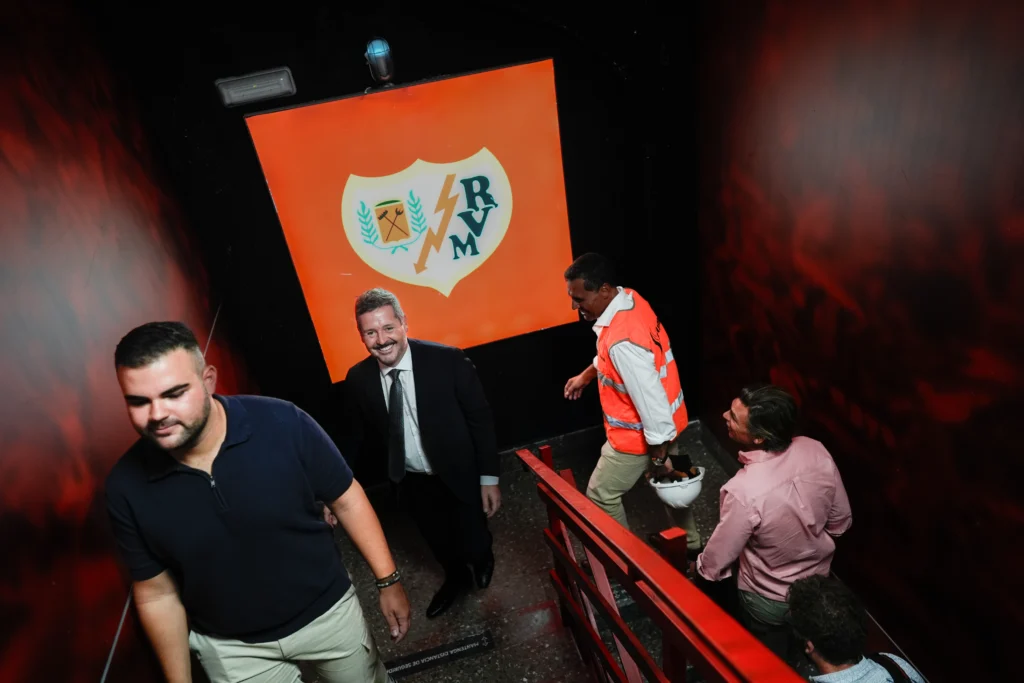 Varias personas suben por una escalera roja con paredes oscuras del Estadio de Vallecas, pasando por una pared con un emblema naranja y blanco del Rayo Vallecano. Un hombre trajeado sonríe a la cámara, mientras otro, con chaleco naranja, lleva un casco, reflejo de la renovación en curso.