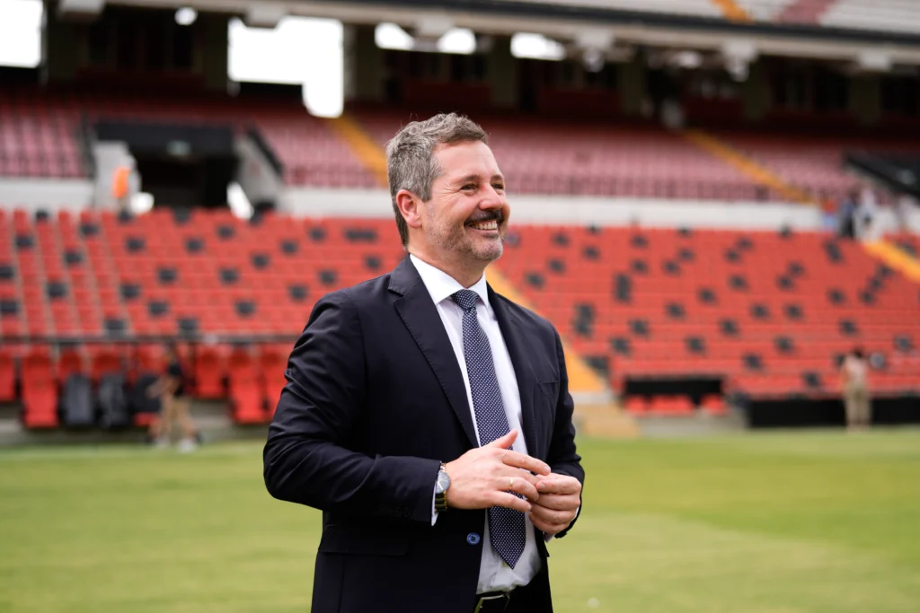 Un hombre con traje y corbata sonríe en un campo de deportes, con los asientos rojos vacíos del Estadio de Vallecas visibles al fondo.