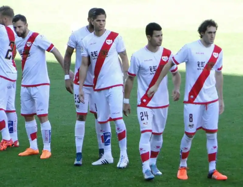 Un grupo de jugadores de fútbol con uniformes blancos con una franja diagonal roja, uniforme del Rayo Vallecano, de pie sobre un campo de hierba. En primer plano se ven cuatro jugadores, algunos mirando a los lados y otros con los brazos a los lados. Abonos 2025/2026.
