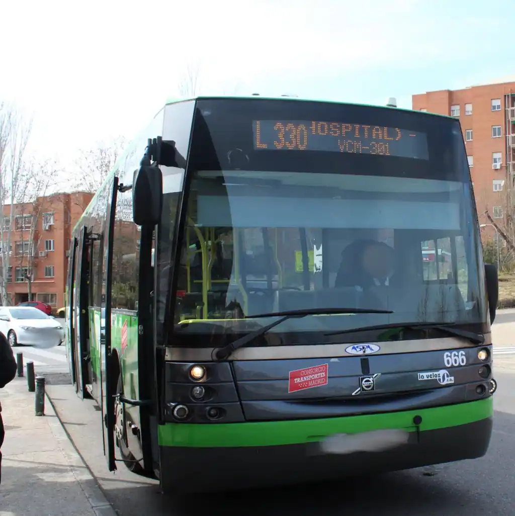 Un autobús urbano verde y negro con la etiqueta Hospital L330 está aparcado en una parada de autobús en una calle de la ciudad, donde frecuencias de los autobuses ayudan a los pasajeros a planificar sus viajes en medio de edificios de apartamentos de ladrillo y algunos coches en el fondo. Rivas recibirá 93.000 euros en ayudas al transporte público