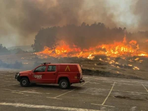 Un vehículo rojo de respuesta a emergencias está aparcado en un terreno vacío mientras las grandes llamas y el denso humo de los incendios arden entre la maleza y los árboles del fondo.
