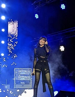Aitana, vestida de negro, actúa en el escenario con una sonrisa, sosteniendo un micrófono mientras las luces azules del escenario y el humo crean una atmósfera eléctrica de fondo. Aitana, vestida de negro, actúa en el escenario con una sonrisa, sosteniendo un micrófono mientras las luces azules del escenario y el humo crean una atmósfera eléctrica de fondo.