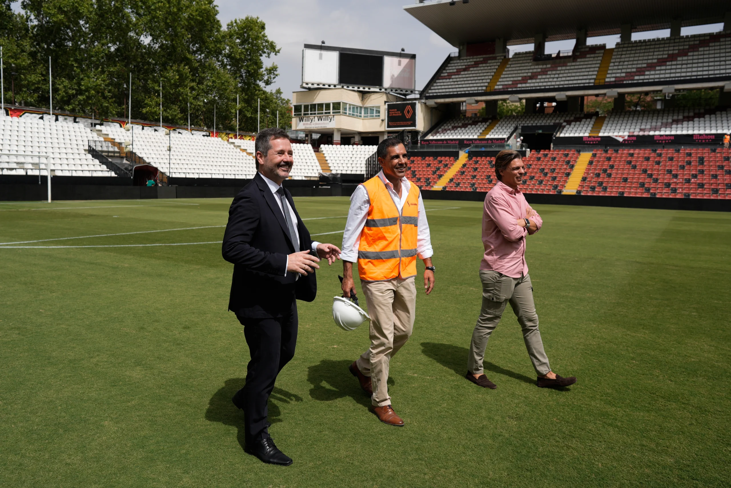 Tres hombres caminan por el campo de fútbol durante la renovación del Estadio de Vallecas en la Comunidad de Madrid; uno lleva un traje negro, otro un chaleco reflectante de seguridad con un casco y otro una camiseta rosa. Al fondo, los asientos del estadio están casi vacíos.