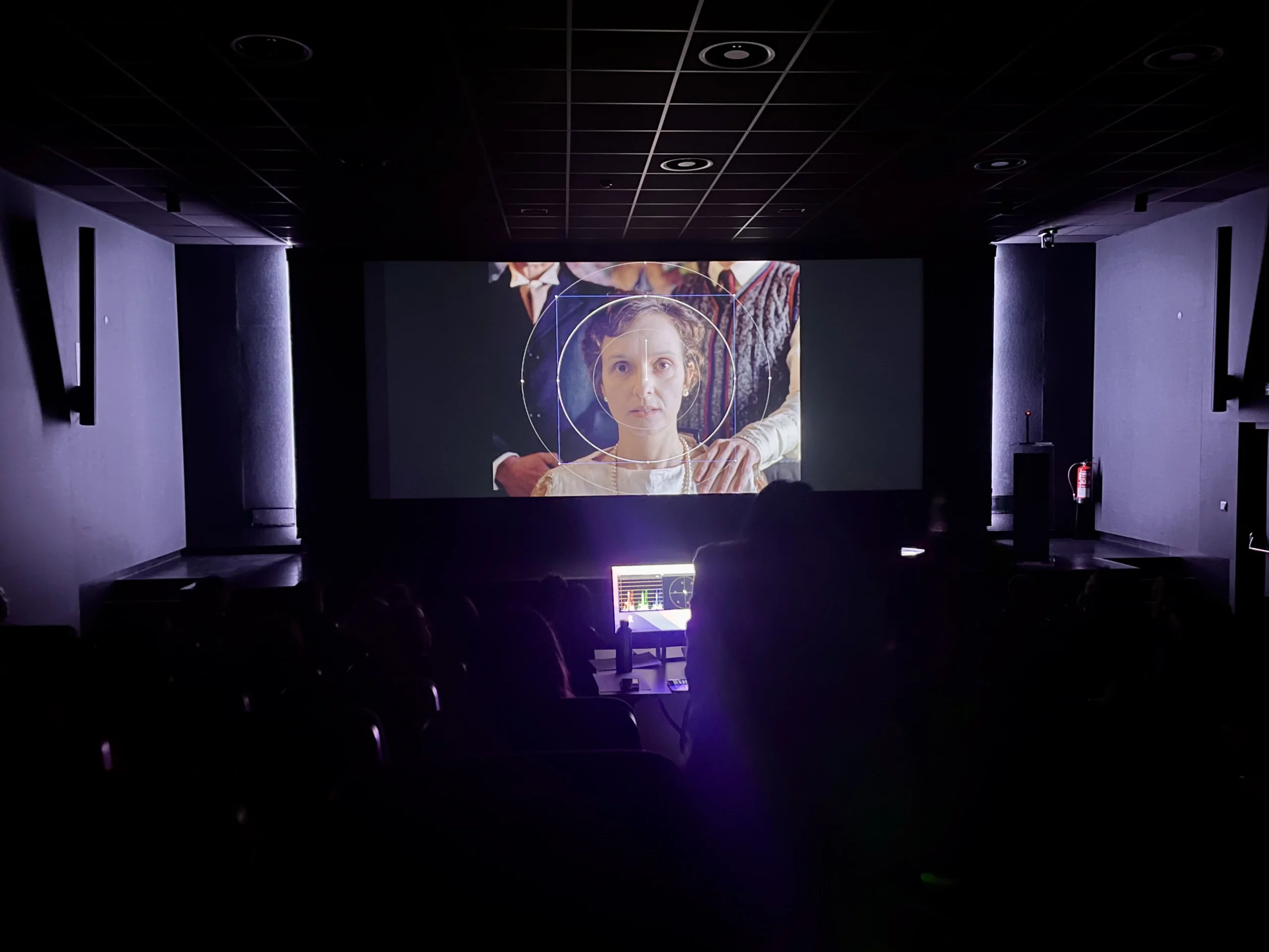 Una sala de cine poco iluminada de la Escuela de Cinematografía de la Comunidad de Madrid proyecta una película en la que aparece una mujer vestida de época. El público está sentado en la sombra, con la luz de un panel de control que ilumina parte de la sala.
