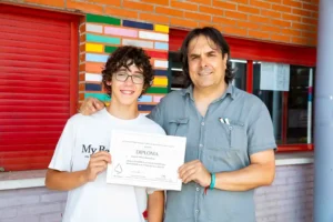 Un estudiante sonriente sostiene un diploma de la Olimpiada Matemática Junior de Madrid mientras está de pie junto a un adulto, posiblemente un profesor, que tiene un brazo alrededor del hombro del estudiante. Están delante de una pared de ladrillo rojo con azulejos de colores.