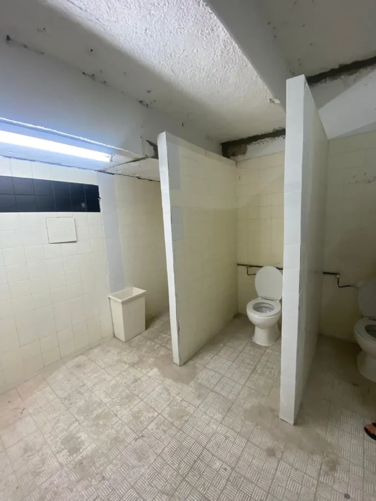 Un aseo público con dos inodoros separados por paredes parciales -sin puertas-, superficies alicatadas, una lámpara fluorescente en el techo y una pequeña papelera blanca en una esquina, que recuerda a las instalaciones de la renovación del Estadio de Vallecas.