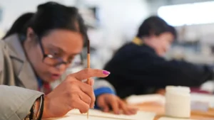 Una persona con un pincel parece estar pintando sobre papel, mientras otra trabaja en una mesa de un aula o estudio de arte. Los materiales artísticos son visibles, lo que refleja cómo los talleres creativos pueden ser beneficiosos para las personas con discapacidad en la Comunidad de Madrid.