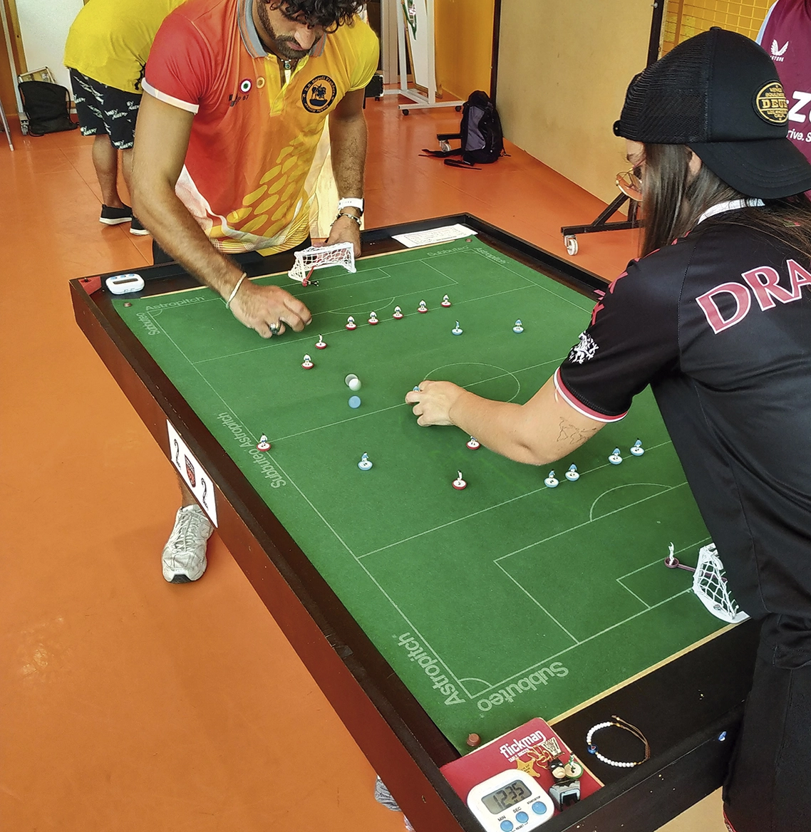 Dos personas juegan a un juego de fútbol de mesa llamado Subbuteo en un tablero de campo verde. El jugador de la izquierda lleva una camiseta naranja, mientras que el de la derecha lleva una camiseta y una gorra negras. En el campo se colocan pequeñas figuritas.