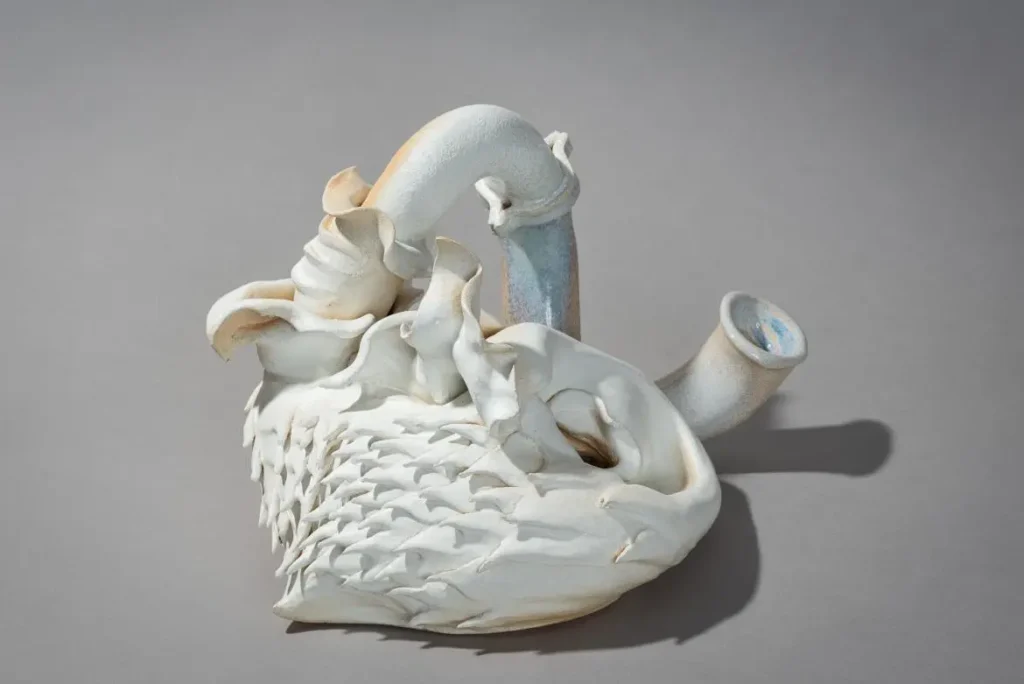Una escultura de cerámica blanca que se asemeja a una forma abstracta y orgánica con tubos curvados y patrones texturizados similares a escamas, presentada en exposiciones y exhibida sobre un fondo gris liso.