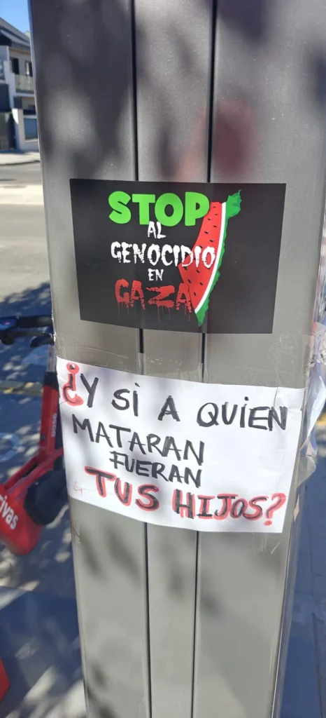 Un poste metálico con dos carteles en español; el de arriba dice Stop al genocidio en Gaza con una ilustración de una sandía y banderas palestinas, mientras que el de abajo dice ¿Y si a quién mataran fueran tus hijos? en texto negro y rojo en negrita.