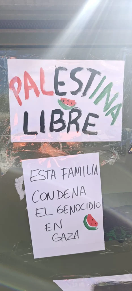 Dos carteles hechos a mano están pegados a una superficie. El cartel superior dice PALESTINA LIBRE con ilustraciones de sandías, parecidas a banderas palestinas. El de abajo dice ESTA FAMILIA CONDENA EL GENOCIDIO EN GAZA, creado por madres y padres. La luz del sol ilumina ambos.