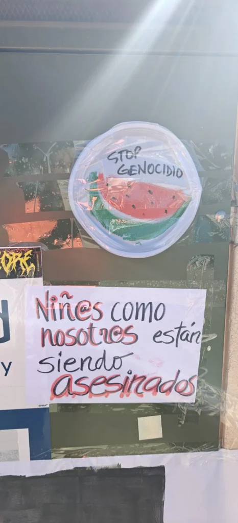 Un bordado circular con una rodaja de sandía y las palabras STOP GENOCIDIO aparece sobre un cartel escrito a mano en español que dice: Niñes como nosotros están siendo asesinados, junto a banderas palestinas creadas por madres y padres del CEIPSO La Luna.