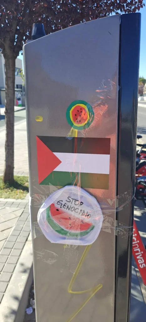 Un poste metálico muestra banderas palestinas, una pegatina circular de una sandía y un papel con una ilustración de una sandía y las palabras Stop Genocide, todo ello pegado con cinta adhesiva transparente. Al fondo se ven una acera y una calle.