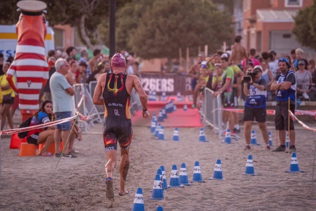Un triatleta que compite en la Liga Nacional de Triatlón lleva bañador, gorro de natación y el número 10 a la espalda mientras corre por un camino de arena bordeado de conos azules, animado por los espectadores y el personal del evento en este emocionante triatlón.