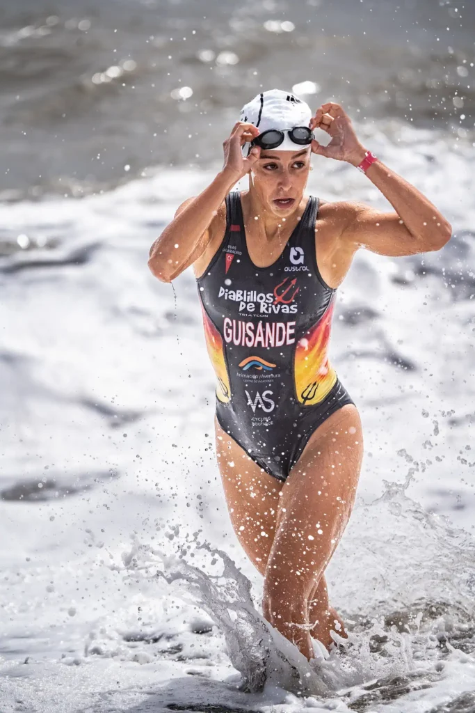 Una atleta con un bañador negro y rojo de Diablillos de Rivas y un gorro de natación sale del agua, ajustándose las gafas mientras las olas del mar chapotean a su alrededor, orgullosa de representar a su equipo en la Liga Nacional de Triatlón.