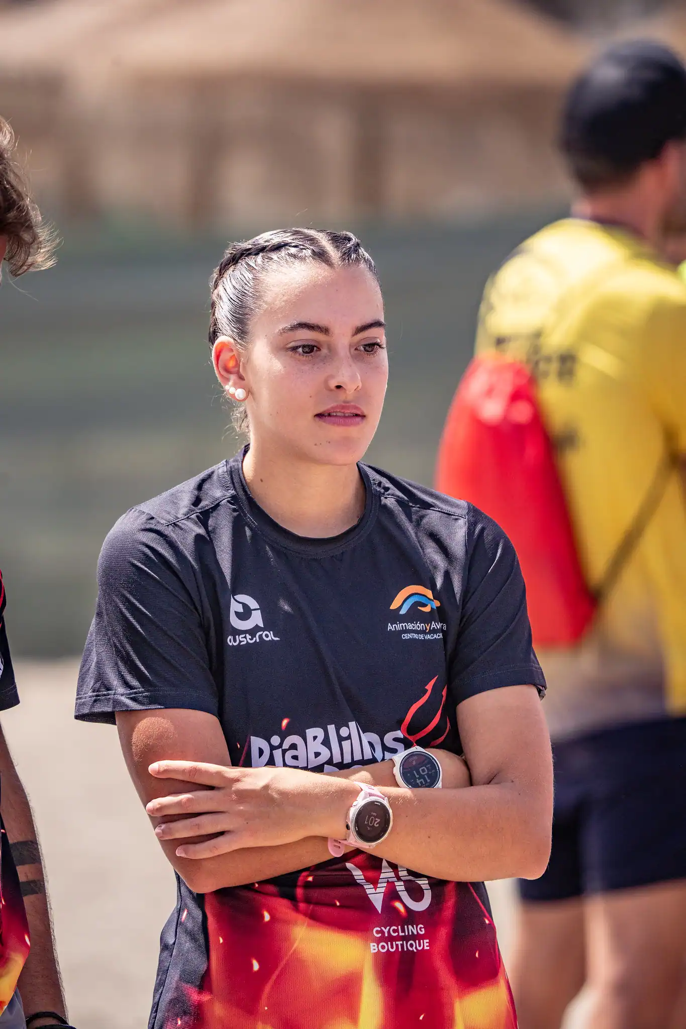 Una mujer joven con el pelo trenzado está de pie con los brazos cruzados, llevando una camiseta deportiva oscura con los logotipos de Diablillos de Rivas y Triatlón, junto con una medalla alrededor del cuello. En el fondo borroso se ven otras personas.