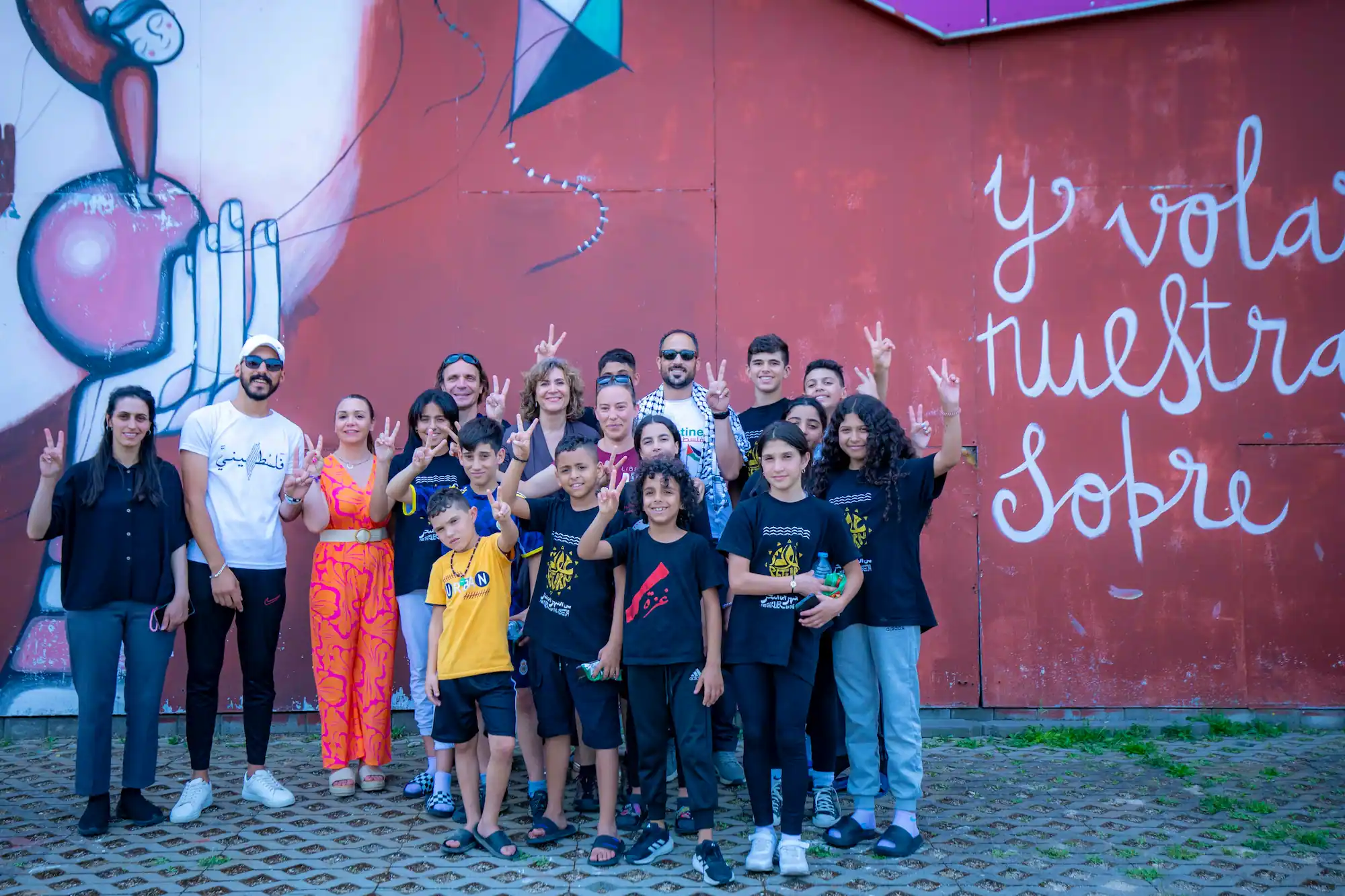 Un grupo de adultos y niños posan juntos frente a un mural en el que se ven grandes manos y las palabras "y volar nuestra" en una pared roja. Muchos de los miembros del grupo, que apoyan a Rivas con Palestina, sostienen carteles de la paz y sonríen.