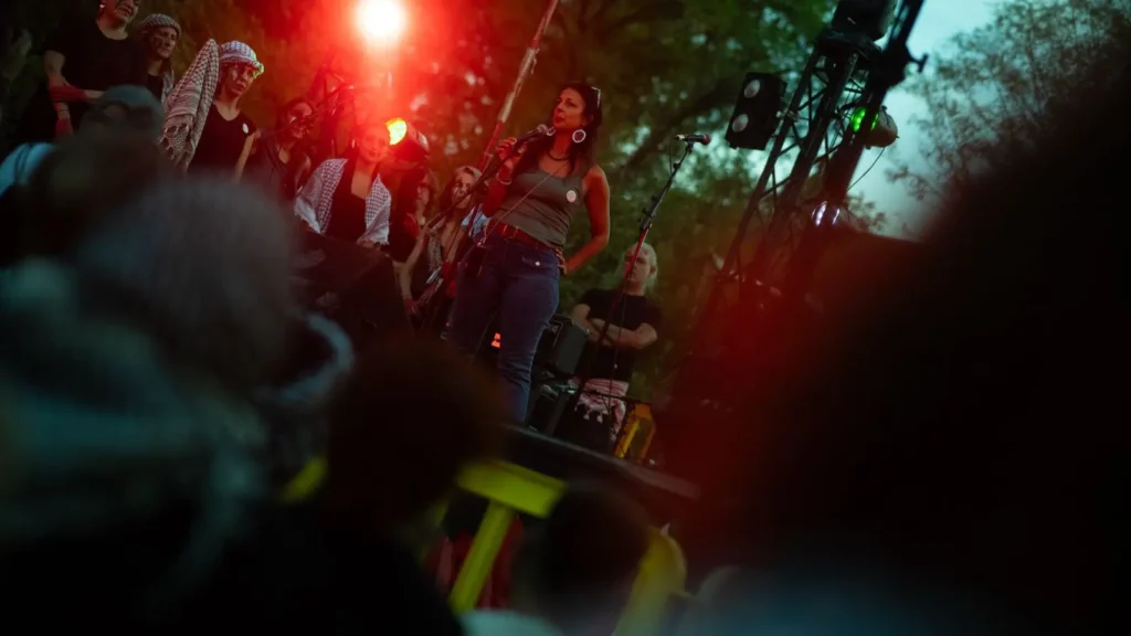 Una mujer habla por un micrófono en un escenario al aire libre, rodeada de varias personas. Las luces rojas del escenario brillan, con árboles y equipos de escenario visibles en el fondo. El público aparece ligeramente difuminado en primer plano.