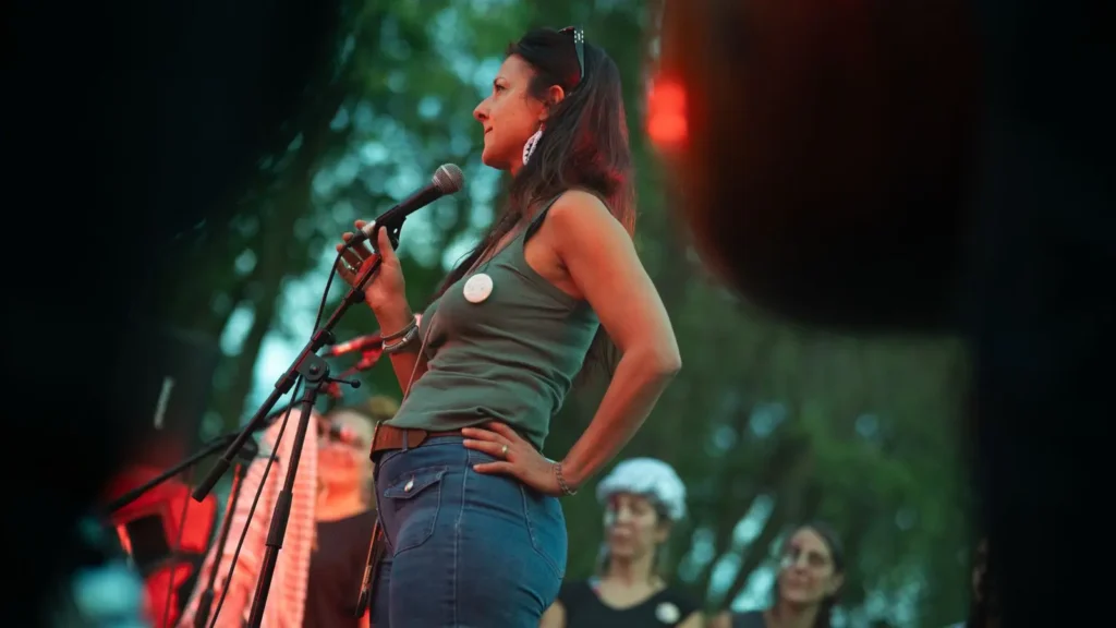 Una mujer está de pie al aire libre sosteniendo un micrófono y hablando, con una mano en la cadera. Está de cara al público y al fondo se ven varias personas entre los árboles. La iluminación está teñida de rojo.