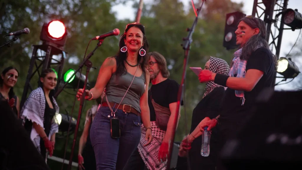 Una mujer frente a un micrófono en un escenario al aire libre, sonríe mientras varias personas están de pie o hablan a su alrededor. Al fondo se ven las luces del escenario y los árboles.