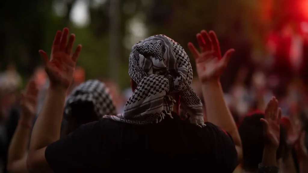 Una persona con camisa negra y un pañuelo estampado en la cabeza levanta ambas manos en el aire entre una multitud de personas al aire libre, con luz roja y natural de fondo.