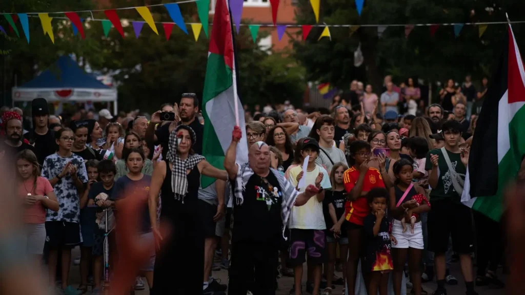 Una gran multitud permanece al aire libre bajo banderas de colores; una persona en el centro sostiene una bandera palestina. Personas de distintas edades y apariencias miran al frente, algunas con expresiones serias.
