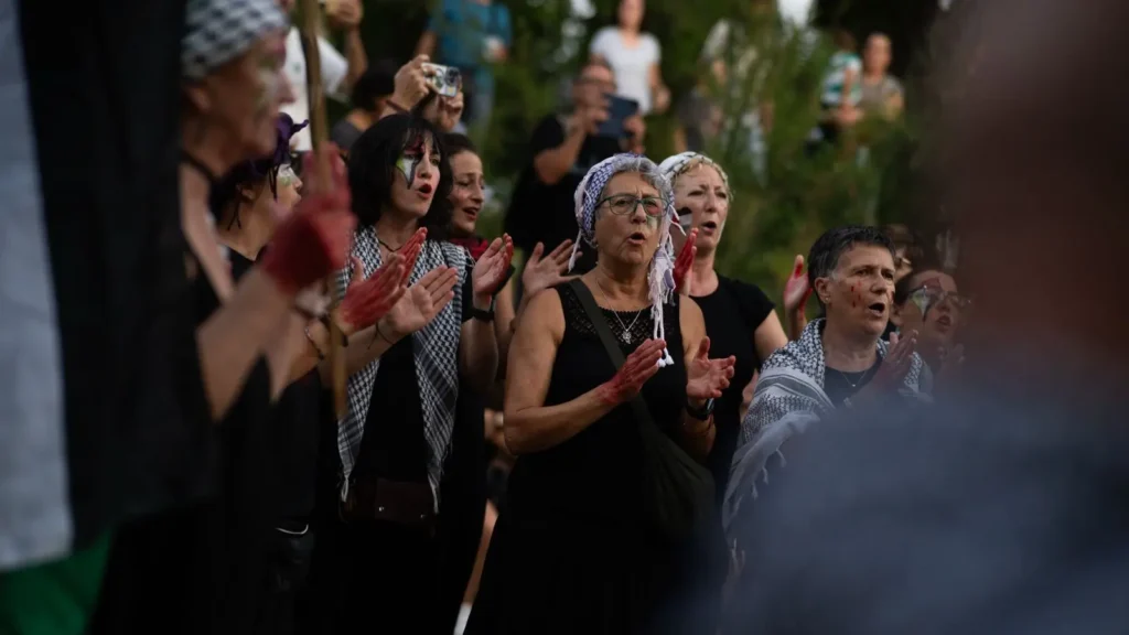 Un grupo de personas vestidas de negro con keffiyehs y pintura roja en las manos aplauden y cantan al aire libre durante una concentración o protesta pública. Al fondo se ven algunos espectadores y árboles.