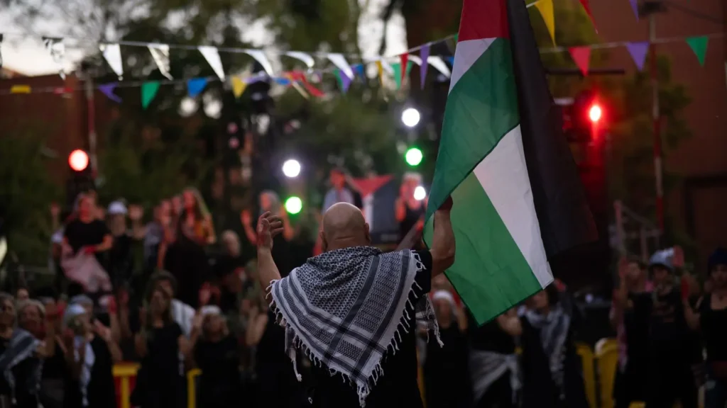 Una persona con un keffiyeh sobre los hombros sostiene una bandera palestina en una reunión al aire libre con banderolas triangulares de colores, una multitud difuminada y luces de fondo.