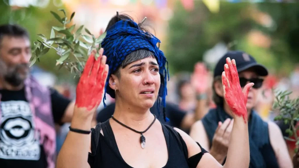Una mujer con un pañuelo azul en la cabeza levanta las manos, cubiertas de pintura roja, mientras parece emocionada en una reunión al aire libre. Al fondo se ve a otras personas.