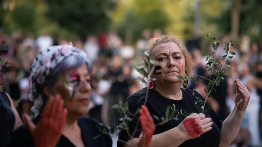 Dos mujeres, de pie entre una multitud al aire libre, sostienen ramas de olivo con las manos y la cara pintadas de rojo y blanco. Ambas llevan camisas negras, y al fondo se ve vegetación y árboles.