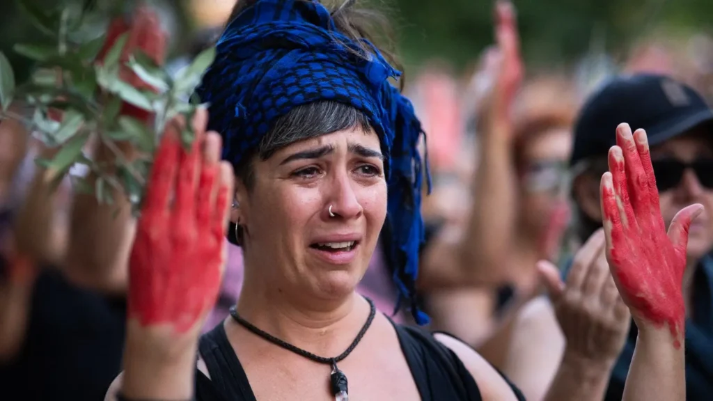 Una persona con un pañuelo azul y negro en la cabeza y pintura roja en las manos, de pie entre la multitud, parece emocionada con lágrimas en la cara. En el fondo se ven otras personas con las manos levantadas.