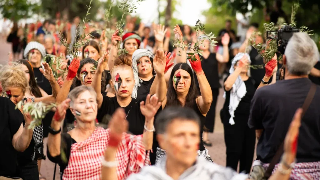 Un grupo de personas participa en una manifestación, muchas con pintura roja en las manos y la cara, algunas con ramas de olivo en las manos y la mayoría vestidas de negro. Permanecen al aire libre con los brazos en alto.