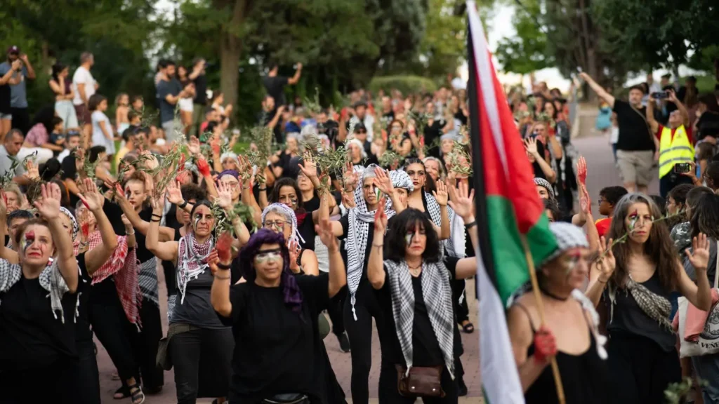 Un numeroso grupo de personas, en su mayoría mujeres, marchan al aire libre sosteniendo ramas de olivo y levantando las manos. Muchas llevan ropa negra y pañuelos blancos y negros, y una persona porta una bandera palestina. Al fondo se ven árboles.