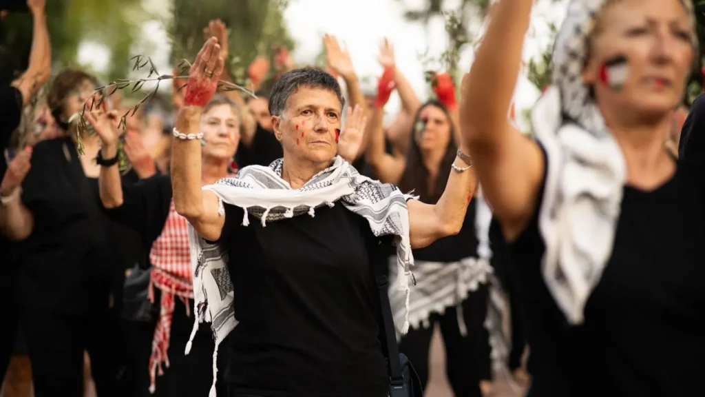 Un grupo de mujeres vestidas de negro y con pañuelos a cuadros levantan los brazos al unísono al aire libre. Algunas tienen las manos y la cara pintadas de rojo; otras sostienen ramas de olivo. El ambiente parece solemne y decidido.