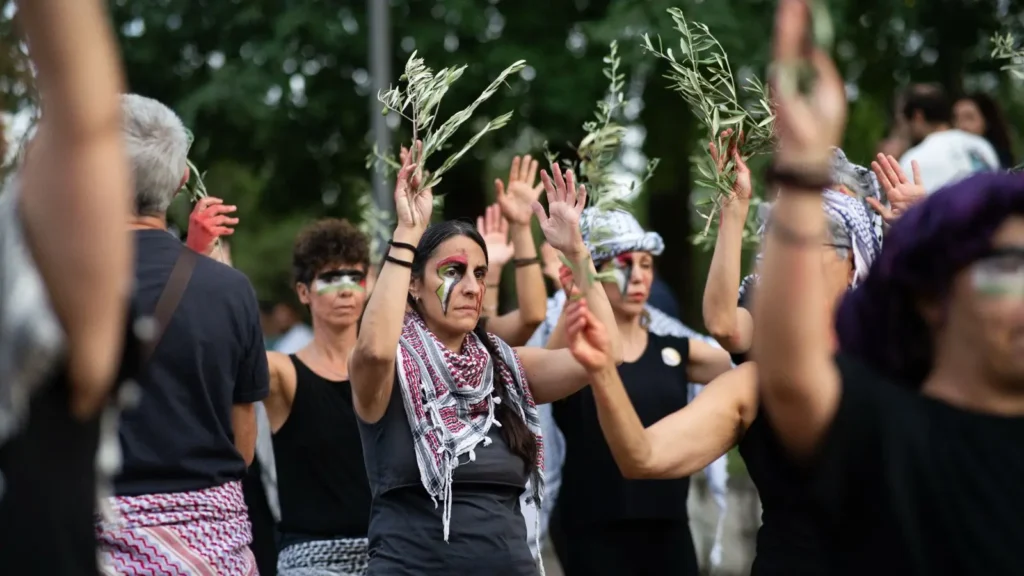 Un grupo de personas permanecen al aire libre con los brazos en alto, sosteniendo ramas de olivo. Muchos llevan ropa negra, pañuelos estampados y la cara pintada. Al fondo se ven árboles y más participantes.