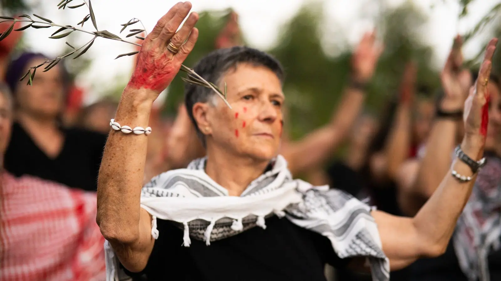 Una persona con un pañuelo estampado y un brazalete de conchas levanta los brazos, con las palmas manchadas de pintura roja, en una reunión al aire libre con otras personas que también han levantado las manos. En el fondo aparecen ramas de árboles y vegetación.