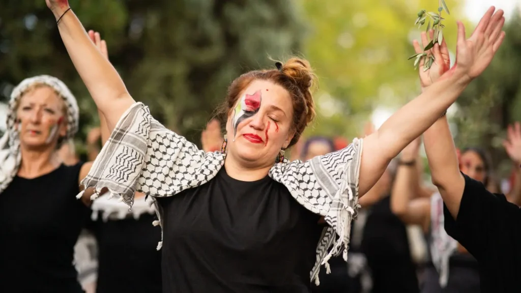 Una mujer con la cara pintada y un pañuelo estampado sobre los hombros está de pie con los brazos levantados al aire libre, los ojos cerrados. Al fondo se ven otras mujeres con atuendos y bufandas similares, también con los brazos en alto.