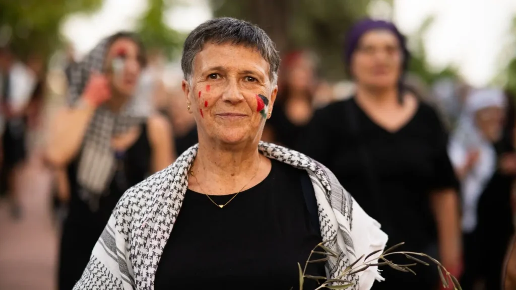Una mujer mayor con el pelo corto y canoso está de pie al aire libre, con una camisa negra y un pañuelo estampado. Tiene la cara pintada de rojo y negro y sostiene unas ramas de olivo. Varias personas aparecen borrosas en el fondo.