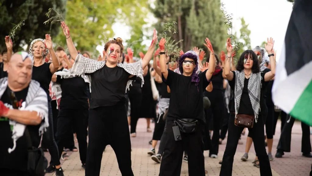 Un grupo de personas vestidas de negro, algunas con keffiyehs y maquillaje que simula heridas, permanecen al aire libre con las manos levantadas durante una manifestación o actuación. Al fondo se ven árboles.