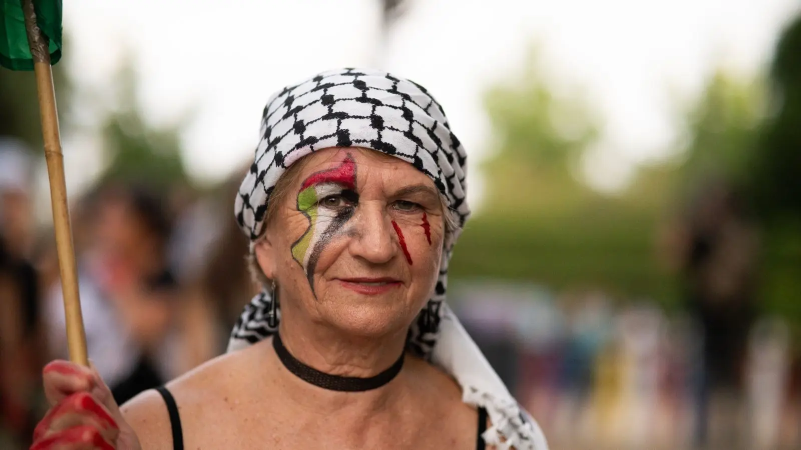 Una mujer mayor con un keffiyeh blanco y negro y una gargantilla está de pie al aire libre. Tiene la cara pintada con los colores de la bandera palestina y sostiene un asta. El fondo es vegetación borrosa. Una mujer mayor con un keffiyeh blanco y negro y una gargantilla está de pie al aire libre. Tiene la cara pintada con los colores de la bandera palestina y sostiene un asta. El fondo es vegetación borrosa.