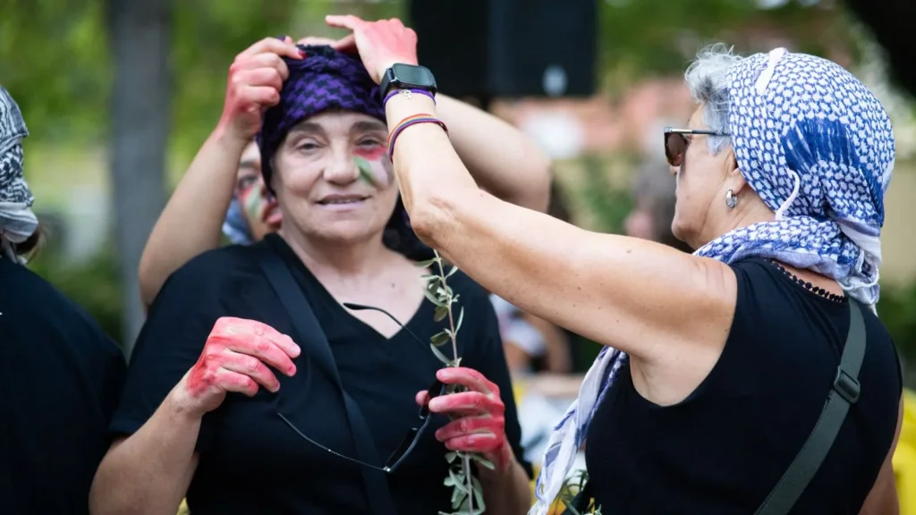 Un grupo de mujeres, algunas con pañuelos estampados, participan en un acto al aire libre. Una mujer ajusta el sombrero de otra y varias llevan las manos pintadas de rojo. Sostienen pequeñas ramas y están rodeadas de vegetación.