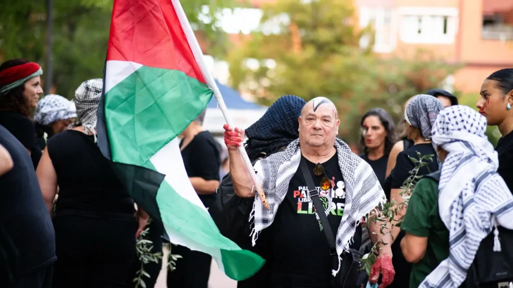 Un grupo de personas, muchas de ellas vestidas de negro y con keffiyehs, permanecen juntas al aire libre. Una persona en el centro sostiene una gran bandera palestina y tiene pintura roja en la mano. Al fondo se ven árboles y edificios.