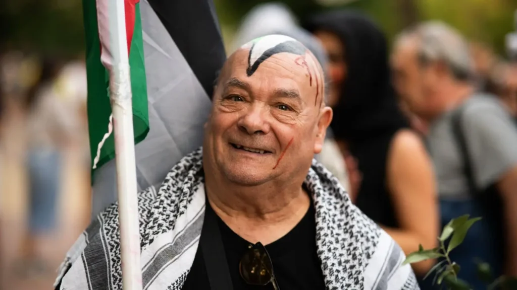 Un hombre sonriente con un pañuelo blanco y negro sobre los hombros está de pie al aire libre, sosteniendo una bandera. Tiene un dibujo negro pintado en la calva. En el fondo borroso se ven otras personas.