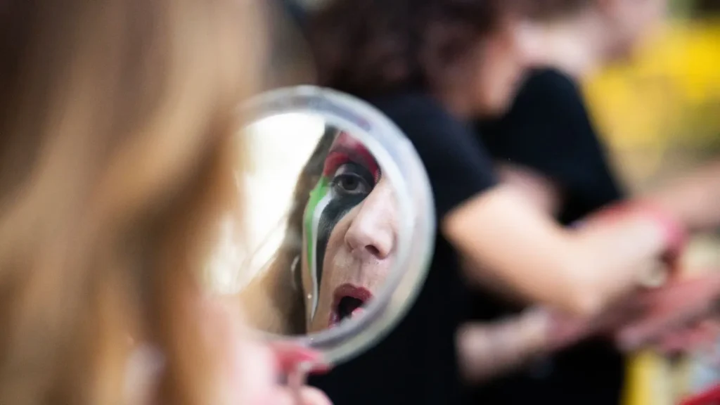 Se ve a una persona con la cara pintada de colores aplicándose maquillaje, reflejada en un espejo de mano redondo, mientras otras personas aparecen difuminadas en el fondo.