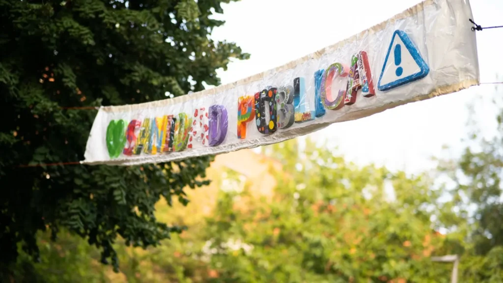 Una pancarta hecha a mano con letras coloridas y decoradas que deletrean SANIDAD PÚBLICA cuelga entre dos árboles en las Fiestas Solidarias de La Pablo 2025.