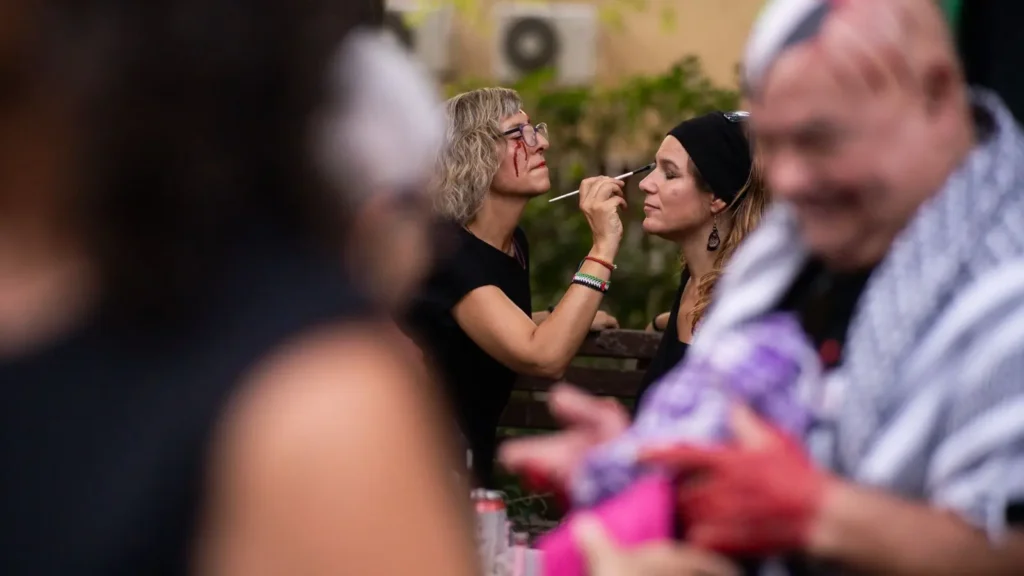 Una mujer maquilla a otra sentada en un banco al aire libre. Se ven otras personas en primer plano, algunas desenfocadas. El escenario parece ser una reunión social informal.