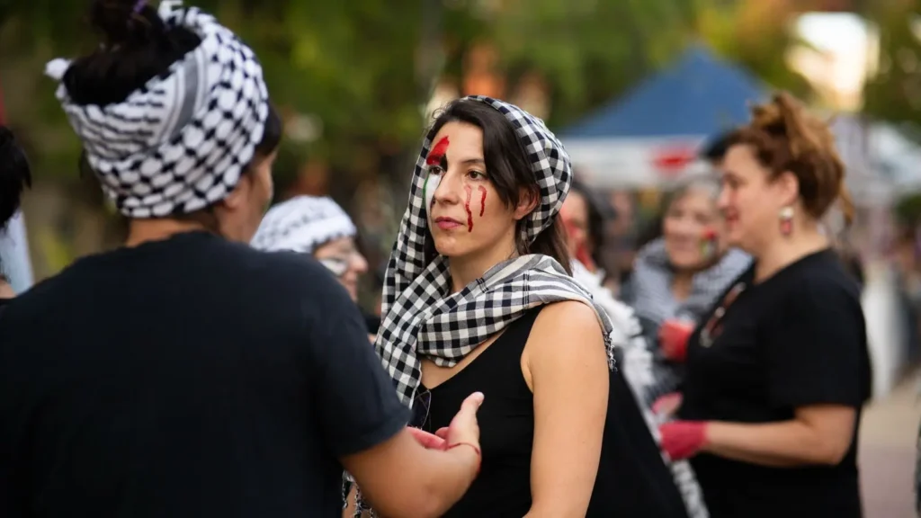 Un grupo de mujeres vestidas de negro y con bufandas a cuadros blancos y negros permanecen al aire libre. Una mujer con pintura roja que parece sangre en la cara mira a otra que le habla. Al fondo se ven árboles y tiendas de campaña.