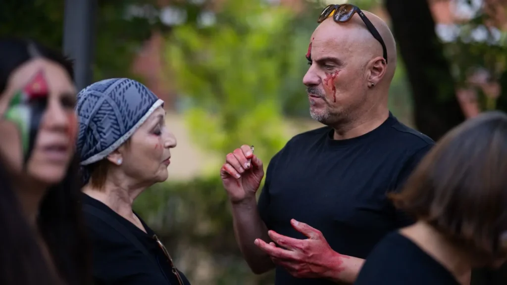Varias personas con maquillaje teatral que parece sangre y heridas están en el exterior. Un hombre maquillado con sangre en la cara y las manos habla con una mujer mayor que lleva un pañuelo en la cabeza. El fondo es vegetación borrosa.