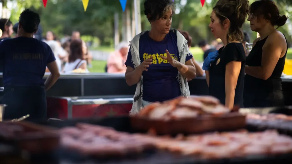 Varias personas conversan de pie cerca de una parrilla al aire libre con bandejas de comida cocinada. Una persona lleva una camiseta que dice fiestas La Paz. Hay banderas de colores al fondo y árboles visibles detrás del grupo.